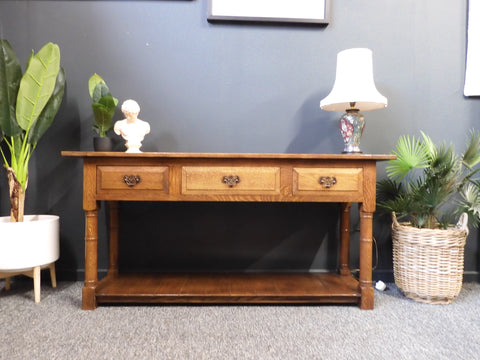 British Arts & Crafts solid oak sideboard or hall table with three drawers, turned legs, and lower shelf, set against a dark grey wall with vibrant artwork.
