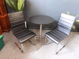 Mid-Century Modern bistro table with speckled granite top and chrome base, flanked by two black ribbed faux leather cantilever chairs on a grey carpet.