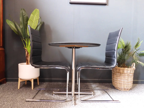 Mid-Century Modern bistro table with speckled granite top and chrome base, flanked by two black ribbed faux leather cantilever chairs on a grey carpet.