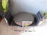 Mid-Century Modern bistro table with speckled granite top and chrome base, flanked by two black ribbed faux leather cantilever chairs on a grey carpet.
