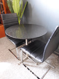 Mid-Century Modern bistro table with speckled granite top and chrome base, flanked by two black ribbed faux leather cantilever chairs on a grey carpet.