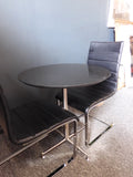 Mid-Century Modern bistro table with speckled granite top and chrome base, flanked by two black ribbed faux leather cantilever chairs on a grey carpet.
