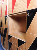 Full view of a tall, artisan-crafted wardrobe from the early 2000s. It features a bold geometric pattern of red, black, and natural birch plywood triangles on its doors, framed by polished metal rounded edges. An open cubby is visible in the center. The base is also metal-clad with cut-out shapes.