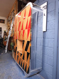 Full view of a tall, artisan-crafted wardrobe from the early 2000s. It features a bold geometric pattern of red, black, and natural birch plywood triangles on its doors, framed by polished metal rounded edges. An open cubby is visible in the center. The base is also metal-clad with cut-out shapes.