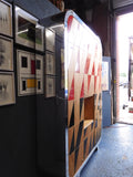 Full view of a tall, artisan-crafted wardrobe from the early 2000s. It features a bold geometric pattern of red, black, and natural birch plywood triangles on its doors, framed by polished metal rounded edges. An open cubby is visible in the center. The base is also metal-clad with cut-out shapes.