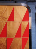 Full view of a tall, artisan-crafted wardrobe from the early 2000s. It features a bold geometric pattern of red, black, and natural birch plywood triangles on its doors, framed by polished metal rounded edges. An open cubby is visible in the center. The base is also metal-clad with cut-out shapes.