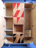 Full view of a tall, artisan-crafted wardrobe from the early 2000s. It features a bold geometric pattern of red, black, and natural birch plywood triangles on its doors, framed by polished metal rounded edges. An open cubby is visible in the center. The base is also metal-clad with cut-out shapes.