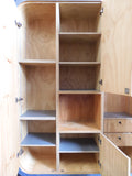 Full view of a tall, artisan-crafted wardrobe from the early 2000s. It features a bold geometric pattern of red, black, and natural birch plywood triangles on its doors, framed by polished metal rounded edges. An open cubby is visible in the center. The base is also metal-clad with cut-out shapes.