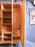 Full view of a tall, artisan-crafted wardrobe from the early 2000s. It features a bold geometric pattern of red, black, and natural birch plywood triangles on its doors, framed by polished metal rounded edges. An open cubby is visible in the center. The base is also metal-clad with cut-out shapes.