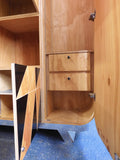 Full view of a tall, artisan-crafted wardrobe from the early 2000s. It features a bold geometric pattern of red, black, and natural birch plywood triangles on its doors, framed by polished metal rounded edges. An open cubby is visible in the center. The base is also metal-clad with cut-out shapes.