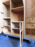 Full view of a tall, artisan-crafted wardrobe from the early 2000s. It features a bold geometric pattern of red, black, and natural birch plywood triangles on its doors, framed by polished metal rounded edges. An open cubby is visible in the center. The base is also metal-clad with cut-out shapes.