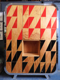 Full view of a tall, artisan-crafted wardrobe from the early 2000s. It features a bold geometric pattern of red, black, and natural birch plywood triangles on its doors, framed by polished metal rounded edges. An open cubby is visible in the center. The base is also metal-clad with cut-out shapes.