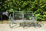 Antique Victorian slatted wrought iron garden bench with scrolled arms in weathered green paint set against a hedge