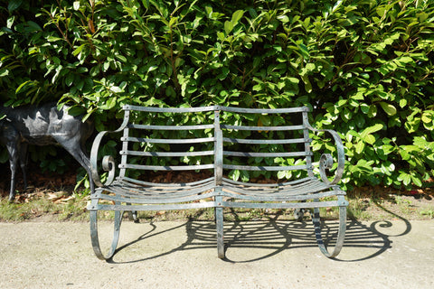 Antique Victorian slatted wrought iron garden bench with scrolled arms in weathered green paint set against a hedge