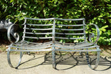 Antique Victorian slatted wrought iron garden bench with scrolled arms in weathered green paint set against a hedge