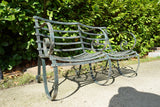 Antique Victorian slatted wrought iron garden bench with scrolled arms in weathered green paint set against a hedge
