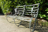 Antique Victorian slatted wrought iron garden bench with scrolled arms in weathered green paint set against a hedge