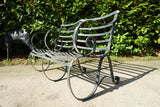 Antique Victorian slatted wrought iron garden bench with scrolled arms in weathered green paint set against a hedge