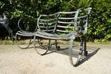 Antique Victorian slatted wrought iron garden bench with scrolled arms in weathered green paint set against a hedge