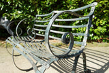 Antique Victorian slatted wrought iron garden bench with scrolled arms in weathered green paint set against a hedge