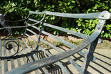 Antique Victorian slatted wrought iron garden bench with scrolled arms in weathered green paint set against a hedge