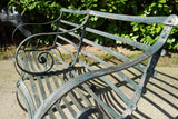 Antique Victorian slatted wrought iron garden bench with scrolled arms in weathered green paint set against a hedge