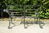 Antique Victorian slatted wrought iron garden bench with scrolled arms in weathered green paint set against a hedge