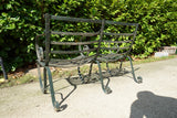 Antique Victorian slatted wrought iron garden bench with scrolled arms in weathered green paint set against a hedge