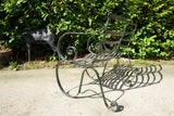 Antique Victorian slatted wrought iron garden bench with scrolled arms in weathered green paint set against a hedge
