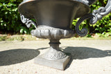 Pair of vintage black cast iron campana garden urn planters with scroll handles and flower medallions on concrete backdrop