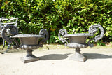 Pair of vintage black cast iron campana garden urn planters with scroll handles and flower medallions on concrete backdrop