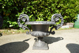Pair of vintage black cast iron campana garden urn planters with scroll handles and flower medallions on concrete backdrop