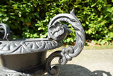 Pair of vintage black cast iron campana garden urn planters with scroll handles and flower medallions on concrete backdrop